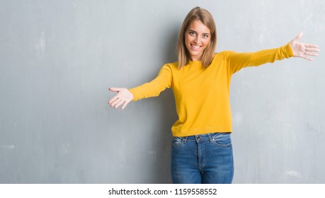 Beautiful young woman standing over grunge grey wall looking at the camera smiling with open arms for hug. Cheerful expression embracing happiness.