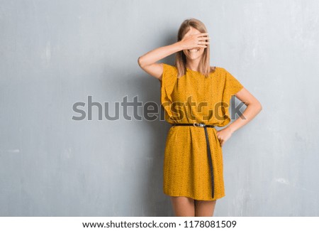 Beautiful young woman standing over grunge grey wall wearing a dress smiling and laughing with hand on face covering eyes for surprise. Blind concept.