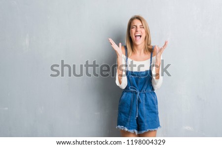 Beautiful young woman standing over grunge grey wall celebrating mad and crazy for success with arms raised and closed eyes screaming excited. Winner concept