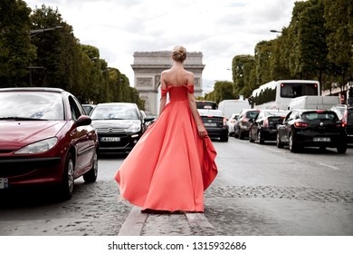 A beautiful young woman stands with her back in a luxurious peach long dress on the middle of the road in Paris, looks at the Arc de Triomphe