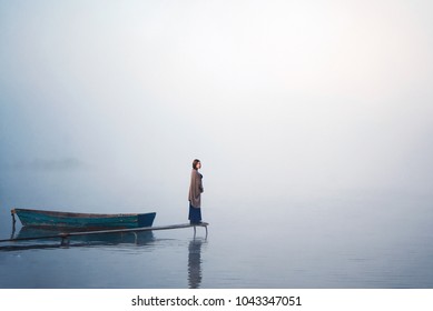 A Beautiful Young Woman Stands In The Middle Of The River In The Fog Near The Boat. Beautiful Background Blur. Fog Background. Solitude, Meditation, Reflection, Thought.