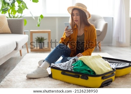 Beautiful young woman staring at smartphone screen in terror, feeling shocked about cancelled flight, sitting near open suitcase with packed clothes at home. Travel ban during covid epidemic