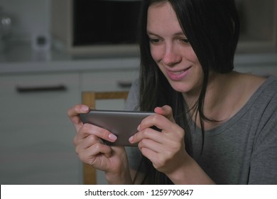 Beautiful Young Woman Stealthily Playing Games On Mobile Phone In The Kitchen At Night.