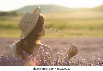 Beautiful young woman with a straw hat holding a bunch of lavender flowers and enjoying their fragrance in the middle of a lanvender field in the light of the setting sun.