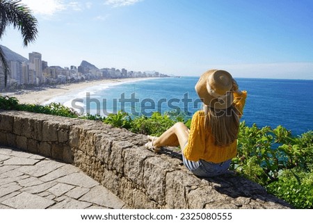 Beautiful young woman with straw hat sitting on wall looking at stunning panoramic view of Rio de Janeiro with Leblon and Ipanema beaches, Rio de Janeiro, Brazil
