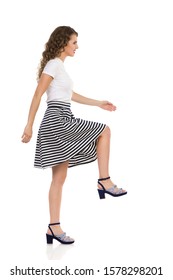 Beautiful Young Woman In Striped, Skirt, High Heels And Blue Top Is Walking Up The Stairs, Smiling, And Looking Away. Side View. Full Length Studio Shot Isolated On White.