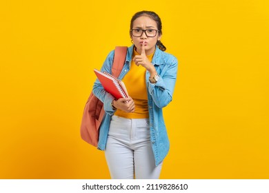 Beautiful young woman student in denim clothes with backpack and holding book, fingers covering lips, making silent gesture on yellow background. Education in high school university college concept