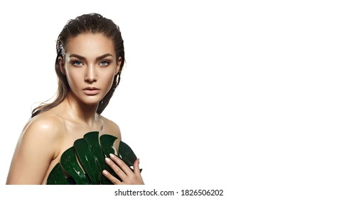 A beautiful young woman in the studio on a white background with wet skin and wet hair holds a large green tropical leaf in her hands and covers a part of her body. Natural cosmetics, natural beauty.
