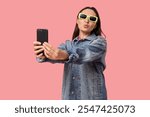 Beautiful young woman in stylish denim jacket and sunglasses taking selfie on pink background