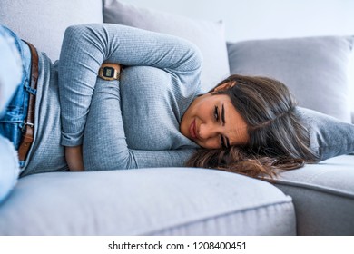 Beautiful young woman suffering from abdominal pain while lying on sofa at home. Girl with stomach ache sitting on bed. Young Hispanic woman sitting in a couch at home and suffering from colics