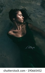 Beautiful Young Woman In A Summer Dress Lying On Her Back In Shallow Water At The Beach With Her Head Turned To The Side And Her Long Black Hair Floating Free