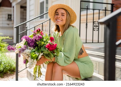 beautiful young woman in summer style outfit smiling happy walking with flowers in city street wearing straw hat fashion trend, sitting on stairs