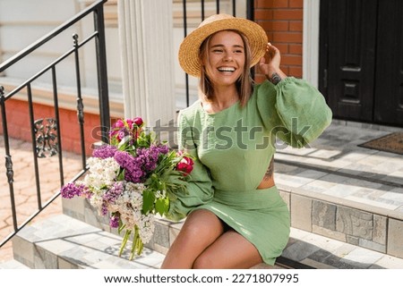 beautiful young woman in summer style outfit smiling happy walking with flowers in city street wearing straw hat fashion trend, sitting on stairs