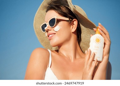 Beautiful Young woman with sun cream on face. Girl holding sunscreen bottle on the beach. Female in hat applying moisturizing lotion on skin.Skin care. Sun protection. Suntan - Powered by Shutterstock