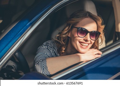 Beautiful Young Woman In Sun Glasses Is Looking At Camera And Smiling While Sitting In A New Car In Car Dealership