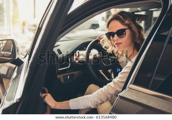 Beautiful young\
woman in sun glasses is looking at camera and smiling while sitting\
in a new car in car\
dealership