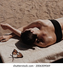 Beautiful young woman sunbathes on the beach. Female sunglasses on beige towel. Minimal aesthetic lifestyle fashion blog, magazine, social media. Sunbathe, relax, chill on summer travel vacations