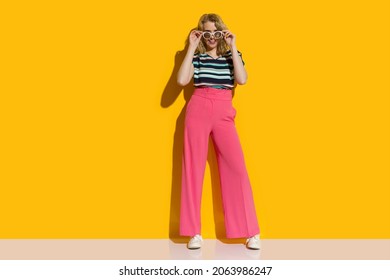 Beautiful Young Woman In Sunglasses, High Waisted Pink Baggy Pants And Striped Top Is Posing In Front Of Yellow Wall. Front View. Full Length Shot.