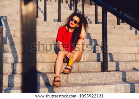 Beautiful young woman in sunglasses sitting on steps, dreaming, holds a bottle of fresh juice, enjoying a sunny day. Outdoors.