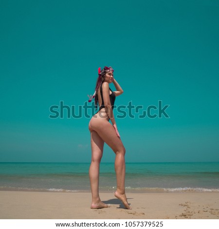 Beautiful young woman in swimsuit with snorkeling standing on the sandy beach over sea and sky background. Pretty girl enjoying sunny summer day on her beach tropical holiday