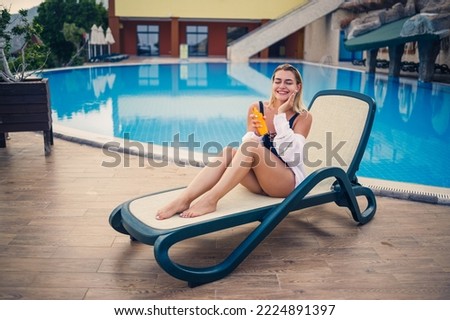 A beautiful young woman in a swimsuit and a white shirt sits on a sun lounger by the pool and rubs her body with sunscreen. Summer skin care, protection against skin burning