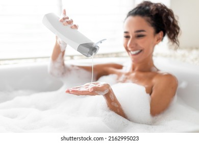 Beautiful Young Woman Taking Bubble Bath, Pouring Shampoo Into Her Hand, Enjoying Home Spa Procedure, Selective Focus. Lovely Millennial Lady Pampering Her Skin, Lying In Bathtub