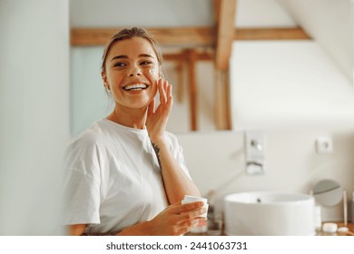 Beautiful young woman taking care of skin by applying moisturizer cream in bathroom - Powered by Shutterstock