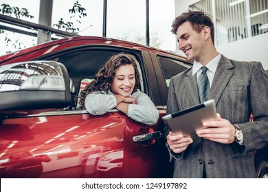 Beautiful Young Woman Is Talking To Handsome Sales Manager While Choosing A Car In Dealership. Man Is Using A Digital Tablet