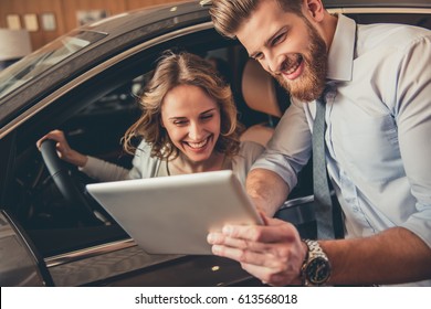 Beautiful Young Woman Is Talking To Handsome Bearded Sales Manager While Choosing A Car In Dealership. Man Is Using A Digital Tablet