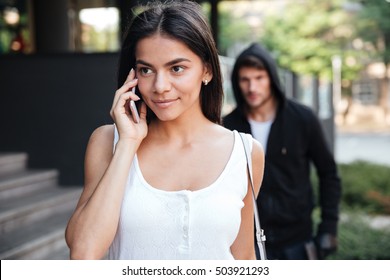 Beautiful young woman talking on mobile phone and being stalked by man criminal on the street