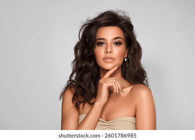 Hermosa mujer joven piel bronceada. Concepto facial y cuidado de la piel del cuerpo Salón. Peinado natural despeinado, cabello largo y ondulado, chica morena, mano en la cara. Aislado sobre fondo gris.