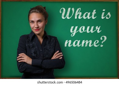 Beautiful Young Woman Teacher (student, Business Woman) In Classical Dress Standing Near A Blackboard With The Inscription What Is Your Name