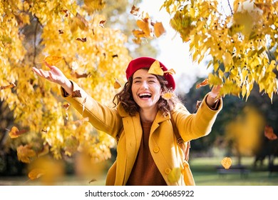 Beautiful young woman throwing autumn yellow leaves. Portrait of joyful woman playing with leaves in park during fall. Happy girl wearing coat and red beret playing with yellow leaves outdoor. - Shutterstock ID 2040685922