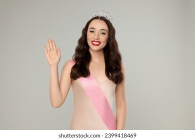 Beautiful young woman with tiara and ribbon in dress on grey background. Beauty contest