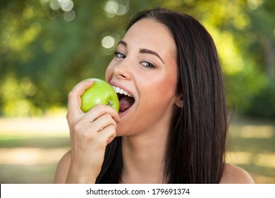 Beautiful young woman with toothy smile biting green fresh apple outdoors
