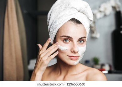 Beautiful young woman with towel wrapped around her head applying face mask at the bathroom