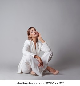 beautiful young woman in a trendy white suit and beige high-heeled shoes, sitting on the floor, a fashion portrait in the studio on a gray background