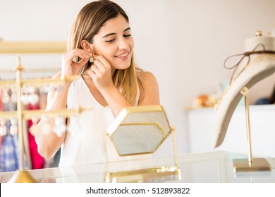 Beautiful young woman trying on a pair of earrings in front of a mirror at a jewelry shop - Powered by Shutterstock