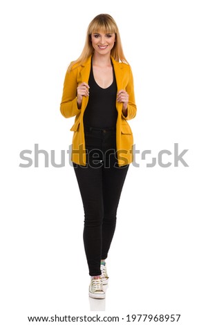 Beautiful young woman in unbuttoned yellow jacket, black jeans and vibrant sneakers is standing looking at camera and smiling. Front view. Full length studio shot isolated on white. Foto d'archivio © 