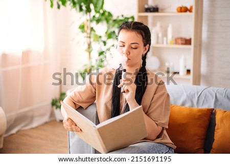 A beautiful young woman uses a disposable electronic cigarette on the couch at home reading a book. vaping concept.