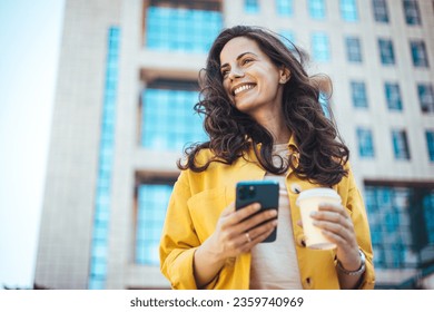 Beautiful young woman is using an app in her smartphone device to send a text message near business buildings. Young positive businesswoman with take away coffee cup in hand using mobile phone  - Powered by Shutterstock