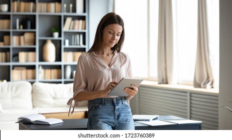 Beautiful young woman using computer tablet, standing in modern living room at home, spending leisure time chatting in social network, writing message, focused blogger freelancer working online