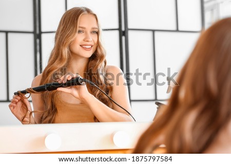 Beautiful young woman using curling iron for hair at home