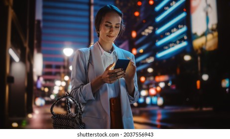 Beautiful Young Woman Using Smartphone Walking Through Night City Street Full of Neon Light. Smiling Thoughtfully Female Using Mobile Phone, Posting Social Media, Online Shopping, Texting. - Powered by Shutterstock