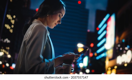 Beautiful Young Woman Using Smartphone Walking Through Night City Street Full Of Neon Light. Portrait Of Gorgeous Smiling Female Using Mobile Phone. Low Angle Shot