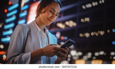 Beautiful Young Woman Using Smartphone Walking Through Night City Street Full of Neon Light. Portrait of Gorgeous Smiling Female Using Mobile Phone. Low Angle Tilt up Shot