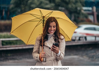 Beautiful Young Woman Using Smartphone And Holding A Yellow Umbrella During A Heavy Rain In The City