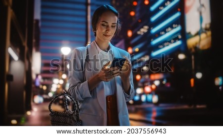 Beautiful Young Woman Using Smartphone Walking Through Night City Street Full of Neon Light. Smiling Thoughtfully Female Using Mobile Phone, Posting Social Media, Online Shopping, Texting.