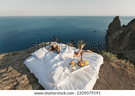 A beautiful young woman wakes up alone in a white bed on a cliff overlooking the sea