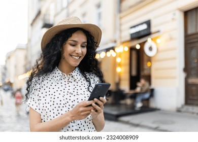 A beautiful young woman walks through the evening city in a hat, a smiling Latin American woman holds a smartphone in her hands. A tourist with curly hair types a message and browses online pages on - Powered by Shutterstock